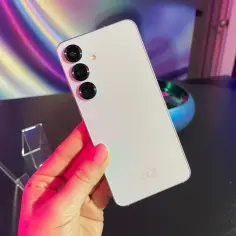 Biały smartfon z trzema aparatami trzymany w dłoni na tle kolorowego, abstrakcyjnego wzoru.